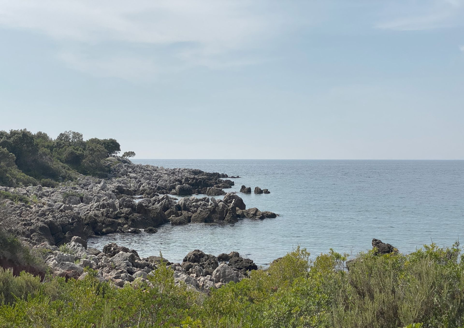 The site today: dark limestone rocks and green scrub running into a calm sea